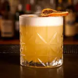 Ein erfrischender Grappa Cocktail mit cremigem Schaum und einer getrockneten Orangenscheibe, serviert in einem Glas mit geometrischem Muster.