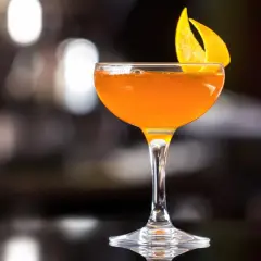 Ein leuchtend oranger Martini Rosso Cocktail, garniert mit einer Orangenzeste, steht auf einer dunklen Oberfläche.