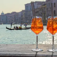 Zwei Aperol Spritz mit Eis und Orangenscheiben stehen auf einer Holzplanke. Im Hintergrund Venedig mit Gondel. Perfekt für Aperol Sekt trocken oder halbtrocken.
