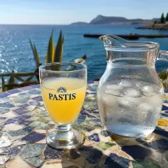 Ein Glas Pastis und ein Krug Eiswasser stehen auf einem bunten Mosaiktisch am Meer. So trinkt man Pastis.