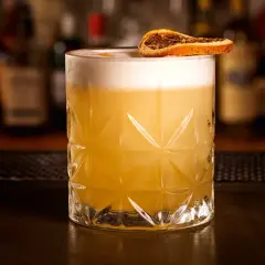 Ein erfrischender Grappa Cocktail mit cremigem Schaum und einer getrockneten Orangenscheibe, serviert in einem Glas mit geometrischem Muster.