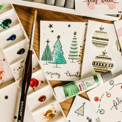 Bunte Aquarellfarben, Pinsel und selbstgemachte Weihnachtskarten mit Bäumen und Kugeln. Perfekt für eine DIY Weihnachtskarte!