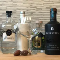 Mehrere Flaschen Gin, darunter Juniper Jack und Bareksten Botanical Gin, stehen neben einem leeren Glas und Zapfen.