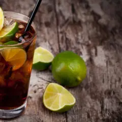 Cuba Libre mit dunklem Rum, Eis und Limettenspalten auf Holzhintergrund.