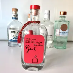 Ein roter Zettel mit der Aufschrift "Und am Ende ergibt alles einen Gin" ist an einer leeren Flasche befestigt. Mehrere Gin-Flaschen stehen im Hintergrund.