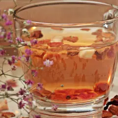Ein Glas **Tee mit Schuss** und getrockneten Früchten, umrahmt von zarten rosa Blüten.