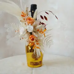 Goldene Weinflasche mit Blumen verpackt. Ein Strauß aus getrockneten Blumen, Rosen und Pampasgras schmückt die Flasche.