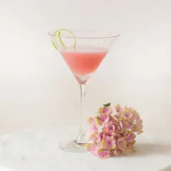Ein rosafarbener Cosmopolitan Cocktail mit Limettenschale und einer Hortensienblüte auf Marmor.