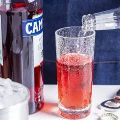 Campari Soda Rezept: Sprudelndes Wasser wird in ein Glas mit Campari gegossen. Eiswürfel und Flaschenöffner liegen bereit.