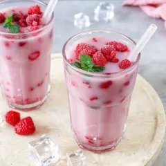 Zwei Gläser mit einem erfrischenden, pinken Drink, gefüllt mit Himbeeren und garniert mit Minze. Eiswürfel und frische Himbeeren liegen daneben.