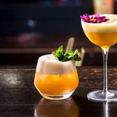 Zwei exquisite Brandy Cocktails, einer mit Minze, der andere mit einer lila Blüte garniert, stehen auf einem dunklen Holztisch.