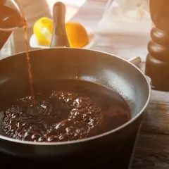 Dunkler Portwein zum Kochen wird in eine Pfanne gegossen, wo er zu köcheln beginnt. Zitrusfrüchte und ein Gewürzstreuer sind im Hintergrund zu sehen.