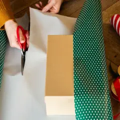 Hände schneiden Geschenkpapier zu, um ein Geburtstagsgeschenk zu verpacken. Rote Schere, Geschenkband und Schleifen liegen bereit.