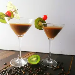 Zwei Martinigläser mit braunem Getränk, garniert mit Kiwi und Kirsche. Kaffeebohnen und eine halbe Kiwi liegen auf dem Tisch. Inspiration für Montenegro Rezepte.