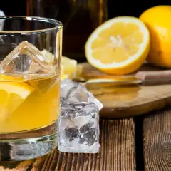 Ein erfrischender Penicillin Cocktail mit Eiswürfeln und Zitronenscheibe auf Holztisch. Daneben liegen frische Zitronen.