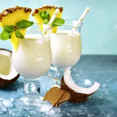 Zwei cremige Malibu Cocktails mit Ananas und Minze, umgeben von Kokosnuss und Eis.
