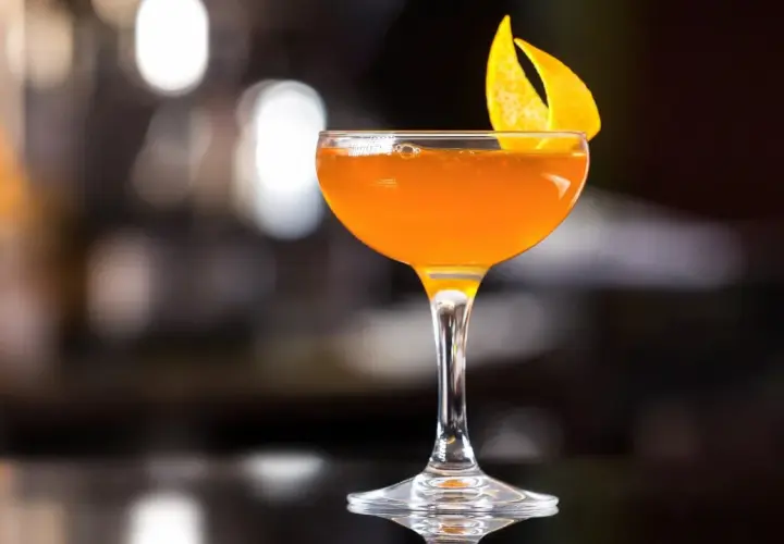 Ein leuchtend oranger Martini Rosso Cocktail, garniert mit einer Orangenzeste, steht auf einer dunklen Oberfläche.