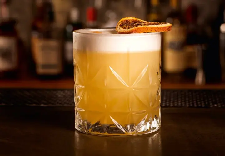 Ein erfrischender Grappa Cocktail mit cremigem Schaum und einer getrockneten Orangenscheibe, serviert in einem Glas mit geometrischem Muster.