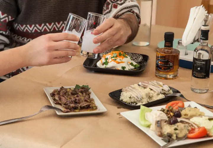 Zwei Gläser mit milchigem Ouzo-Getränk werden angestoßen. Dazu gibt es verschiedene griechische Mezedes und kleine Flaschen Ouzo.