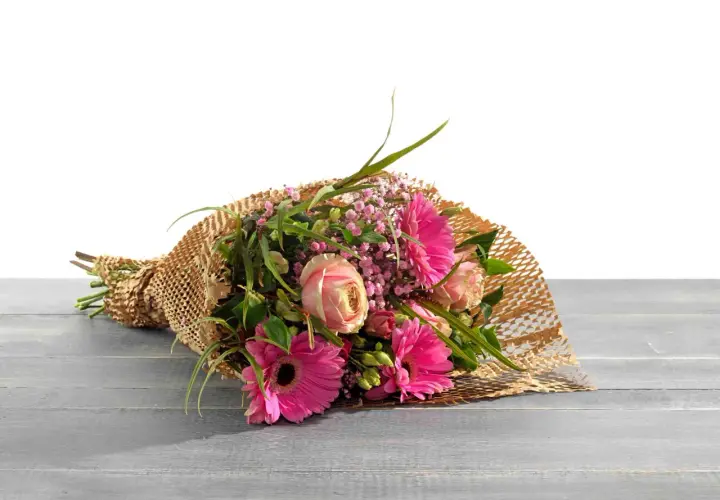 Ein Strauß bunter Blumen, liebevoll als Geschenk verpackt in braunem Papier mit Wabenstruktur, liegt auf einer Holzoberfläche.