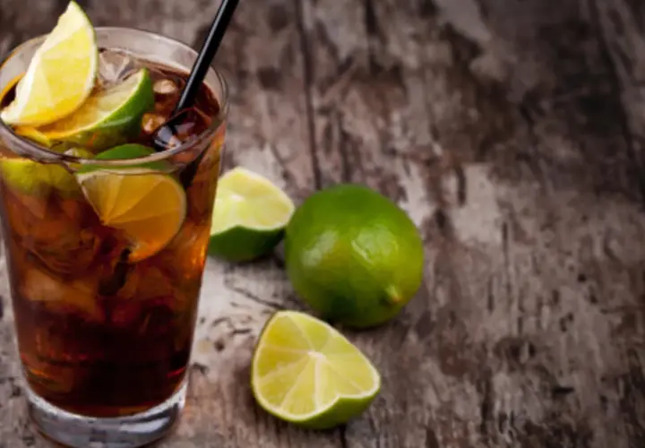 Cuba Libre mit dunklem Rum, Eis und Limettenspalten auf Holzhintergrund.