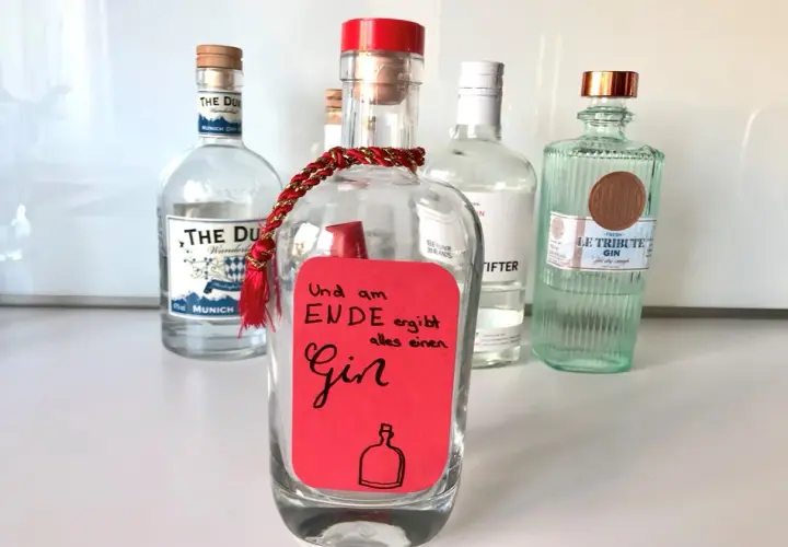 Ein roter Zettel mit der Aufschrift "Und am Ende ergibt alles einen Gin" ist an einer leeren Flasche befestigt. Mehrere Gin-Flaschen stehen im Hintergrund.