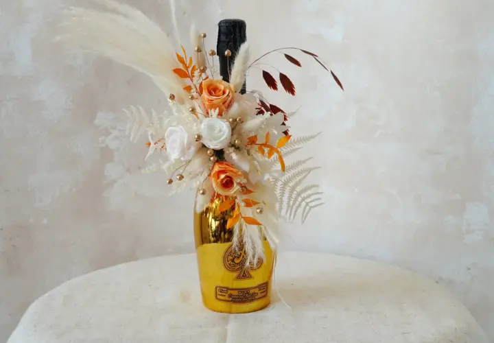 Goldene Weinflasche mit Blumen verpackt. Ein Strauß aus getrockneten Blumen, Rosen und Pampasgras schmückt die Flasche.