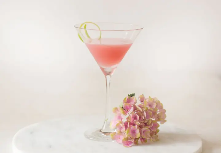 Ein rosafarbener Cosmopolitan Cocktail mit Limettenschale und einer Hortensienblüte auf Marmor.