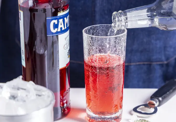 Campari Soda Rezept: Sprudelndes Wasser wird in ein Glas mit Campari gegossen. Eiswürfel und Flaschenöffner liegen bereit.