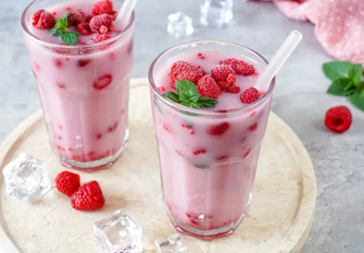 Zwei Gläser mit einem erfrischenden, pinken Drink, gefüllt mit Himbeeren und garniert mit Minze. Eiswürfel und frische Himbeeren liegen daneben.