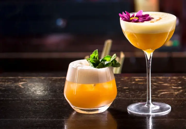 Zwei exquisite Brandy Cocktails, einer mit Minze, der andere mit einer lila Blüte garniert, stehen auf einem dunklen Holztisch.