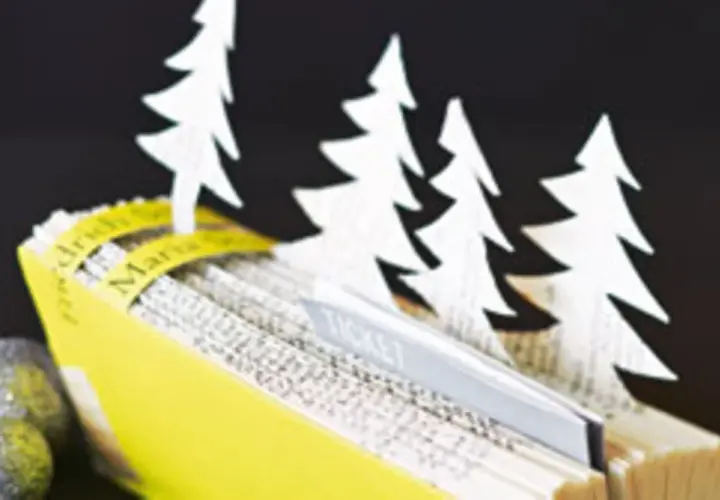 Kreative **Tickets verpacken Ideen**: Ein aufgeschlagenes Buch mit weißen Tannenbäumen und einem Ticket-Einschub, daneben glitzernde Kugeln.