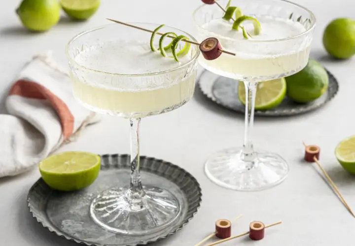 Zwei erfrischende Daiquiri Cocktails mit Limettenschale garniert, umgeben von frischen Limetten.