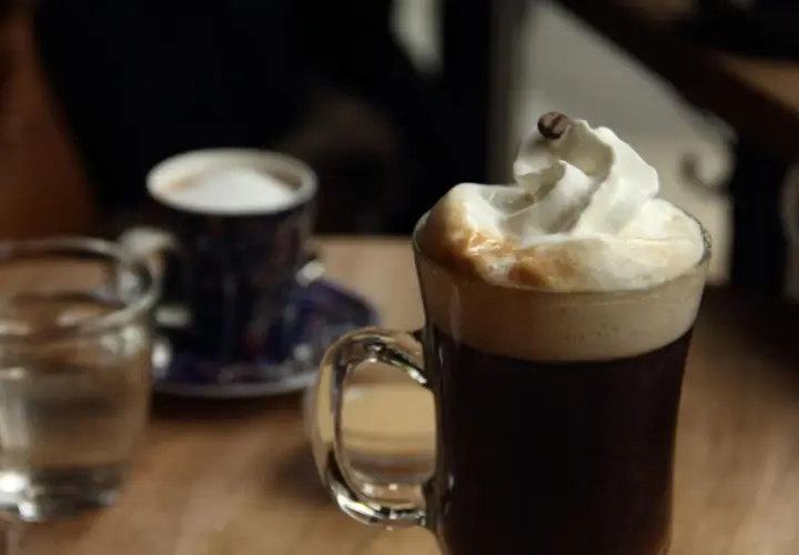 Ein Glas mit Kaffee und Sahne, garniert mit einer Kaffeebohne. Daneben ein kleiner Becher und ein Glas Wasser. Perfekt für einen Kaffee mit Schnaps.