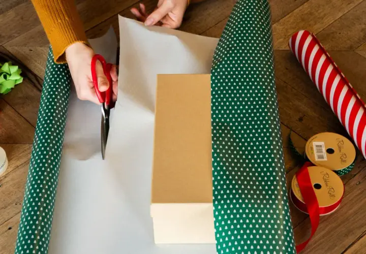 Hände schneiden Geschenkpapier zu, um ein Geburtstagsgeschenk zu verpacken. Rote Schere, Geschenkband und Schleifen liegen bereit.