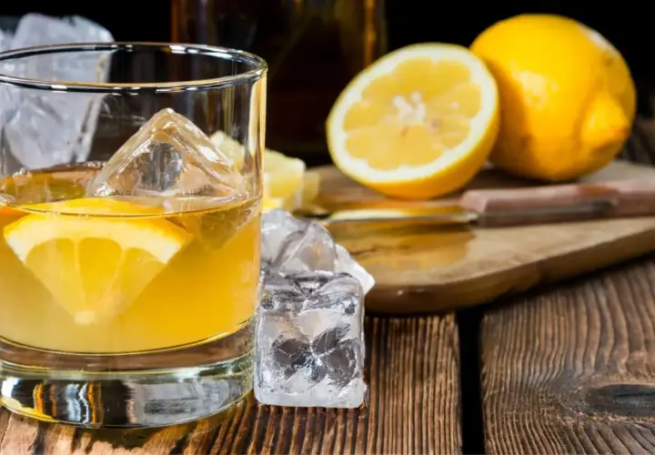 Ein erfrischender Penicillin Cocktail mit Eiswürfeln und Zitronenscheibe auf Holztisch. Daneben liegen frische Zitronen.