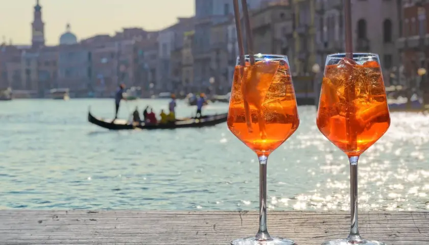 Zwei Aperol Spritz mit Eis und Orangenscheiben stehen auf einer Holzplanke. Im Hintergrund Venedig mit Gondel. Perfekt für Aperol Sekt trocken oder halbtrocken.