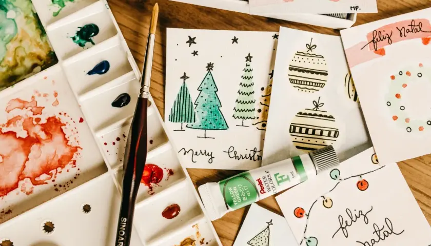 Bunte Aquarellfarben, Pinsel und selbstgemachte Weihnachtskarten mit Bäumen und Kugeln. Perfekt für eine DIY Weihnachtskarte!