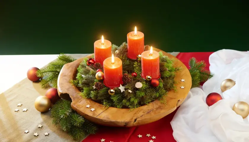 Ein selbstgemachter Adventskranz auf einem Holzteller mit vier brennenden Kerzen, Tannenzweigen, Kugeln und Sternen.