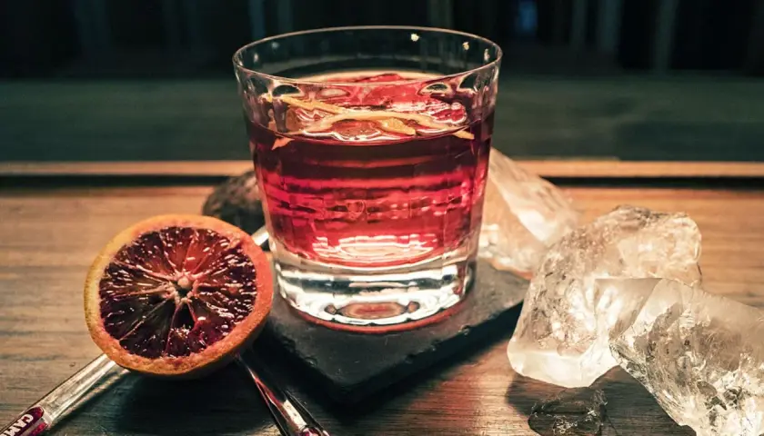 Ein Glas mit roten Cocktails, garniert mit Orangenzesten, neben einer Blutorange und Eiswürfeln auf einem Holztisch.