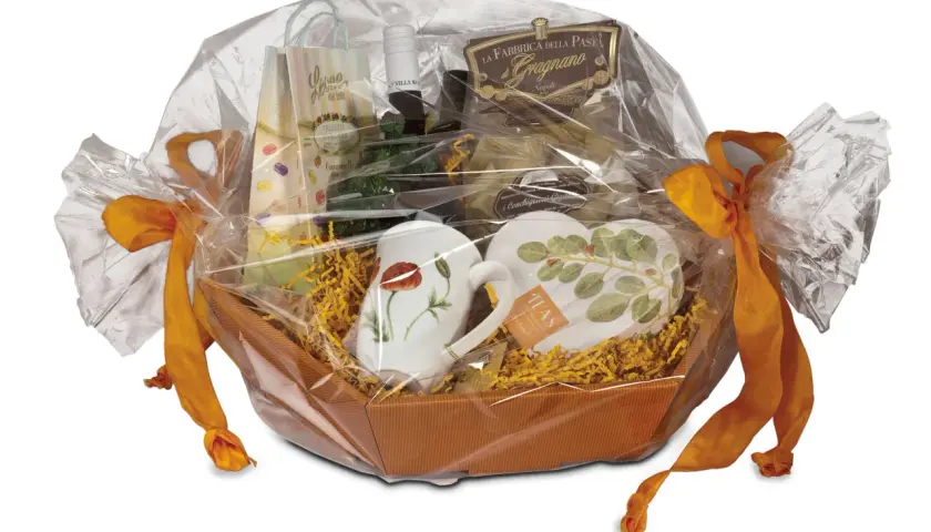 Ein liebevoll zusammengestelltes Geschenk, das mit Folie und orangefarbenen Bändern verpackt ist. Darin befinden sich Tassen, Pasta und weitere Leckereien.