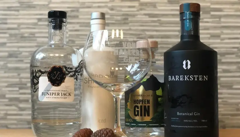 Mehrere Flaschen Gin, darunter Juniper Jack und Bareksten Botanical Gin, stehen neben einem leeren Glas und Zapfen.