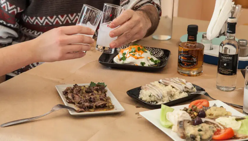 Zwei Gläser mit milchigem Ouzo-Getränk werden angestoßen. Dazu gibt es verschiedene griechische Mezedes und kleine Flaschen Ouzo.