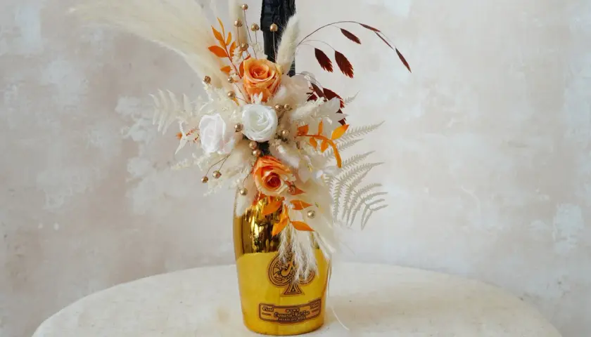 Goldene Weinflasche mit Blumen verpackt. Ein Strauß aus getrockneten Blumen, Rosen und Pampasgras schmückt die Flasche.