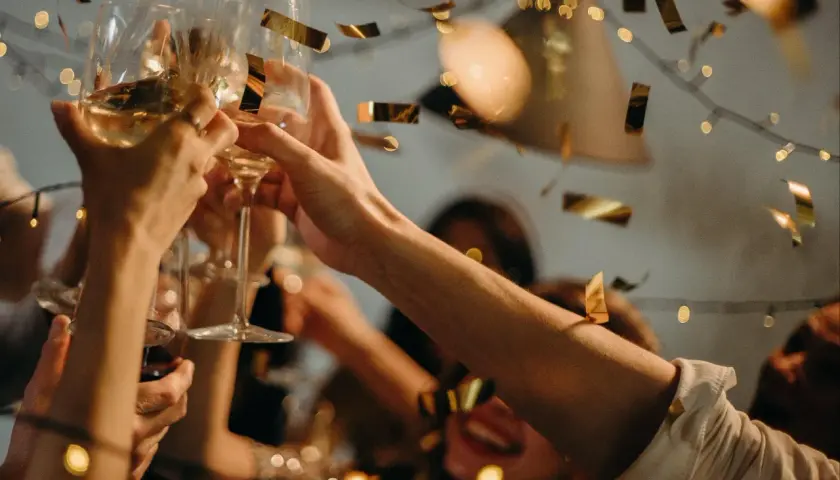 Hände stoßen mit Gläsern an, Konfetti fällt. Ein fröhlicher Anlass, vielleicht ein Jubiläum, wird gefeiert.
