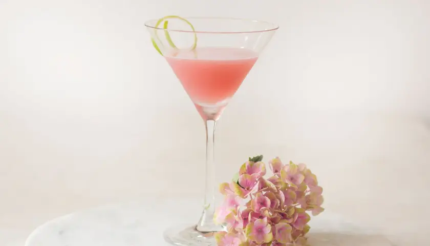 Ein rosafarbener Cosmopolitan Cocktail mit Limettenschale und einer Hortensienblüte auf Marmor.