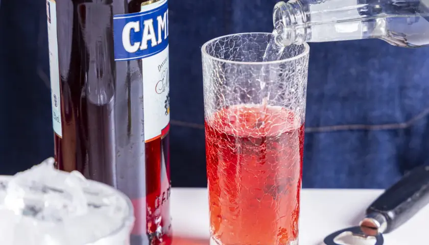 Campari Soda Rezept: Sprudelndes Wasser wird in ein Glas mit Campari gegossen. Eiswürfel und Flaschenöffner liegen bereit.