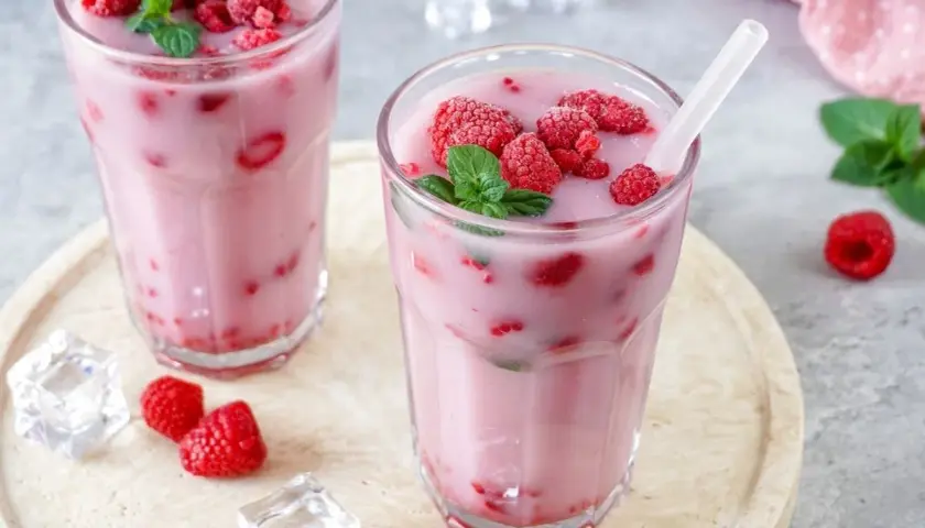 Zwei Gläser mit einem erfrischenden, pinken Drink, gefüllt mit Himbeeren und garniert mit Minze. Eiswürfel und frische Himbeeren liegen daneben.