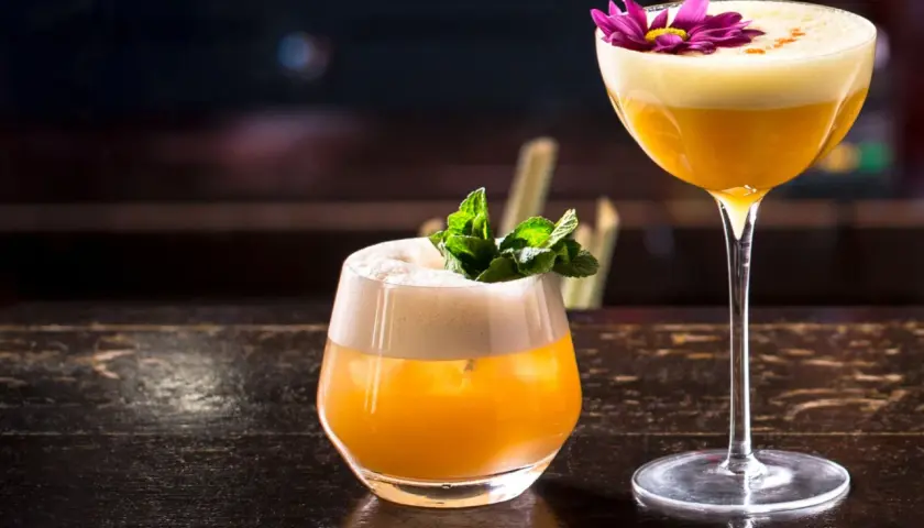 Zwei exquisite Brandy Cocktails, einer mit Minze, der andere mit einer lila Blüte garniert, stehen auf einem dunklen Holztisch.
