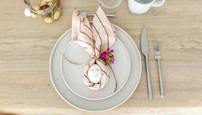 Niedliche Tischdeko Ostern einfach: Ein Ei mit Gesicht, umwickelt von einer Serviette als Hasenohren, mit kleinen Blüten verziert.